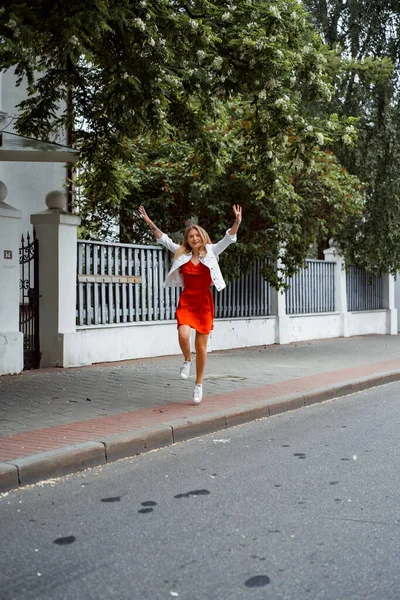 赤いドレスと白いデニムのジャケットを着た美しい若い女性が街を歩き回って幸せな笑顔 — ストック写真