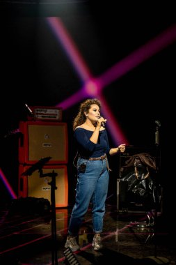 Sahnede genç bir kadın şarkıcı mikrofona şarkı söylüyor..