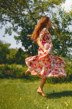 Uzun saçlı, parlak elbiseli genç bir kadın çimenlerde eğleniyor, dans ediyor.