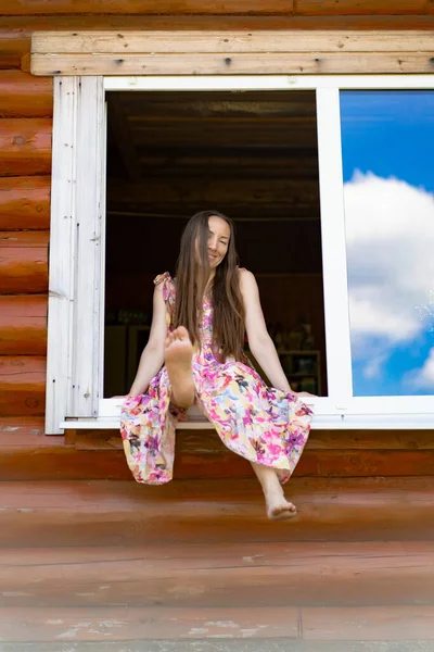 明るい色の太陽の下で長い髪の若い美しい女性が窓に座っています カントリースタイル — ストック写真