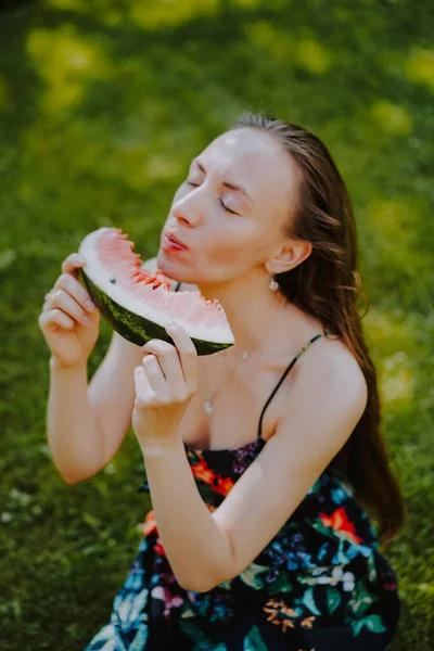 髪の長い美しい若い女性はスイカのスライスと芝生の上にあります 自然の中で熟したスイカを食べる女性 — ストック写真