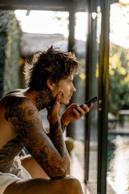 Dövmeli yakışıklı genç adam telefonda hoparlörden konuşuyor.