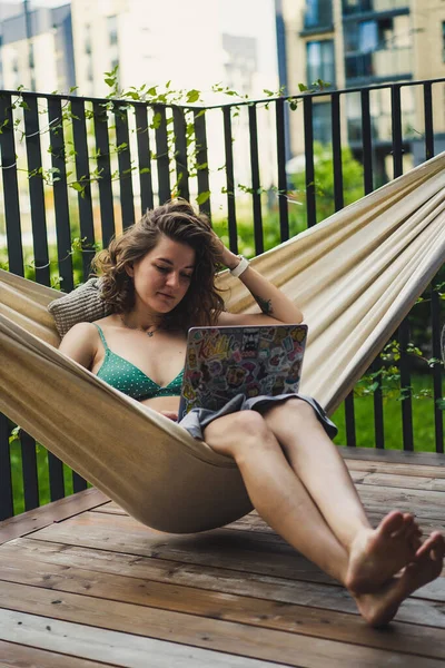 Young woman on a private terrace of a country house in a hammock works on a laptop. Outdoor work, freelancer, programmer, work from home.