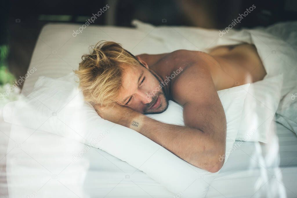 Sexy naked young man is looking at camera while lying in bed. Ca