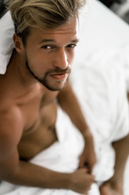Seksi çıplak genç adam yatakta yatarken kameraya bakıyor. Ka