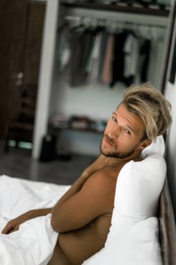 Seksi çıplak genç adam yatakta yatarken kameraya bakıyor. Ka