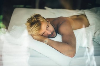 Seksi çıplak genç adam yatakta yatarken kameraya bakıyor. Ka