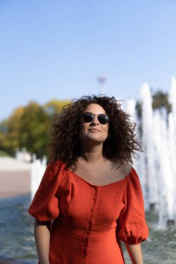 Kıvırcık siyah saçlı, kırmızı elbiseli güzel bir kadın şehir çeşmelerinin yanında yürüyor..