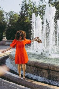 Kıvırcık siyah saçlı, kırmızı elbiseli güzel bir kadın şehir çeşmelerinin yanında yürüyor..