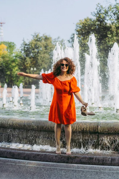 街の噴水の近くの街を歩くと 赤いドレスの中に巻き毛の美しい女性 — ストック写真