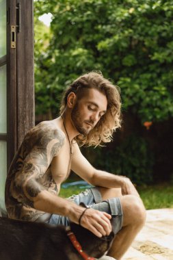 Uzun kıvırcık saçlı ve çıplak gövdeli genç bir adam. Bahçedeki sade bir ortamda bir adam bir köpekle oynuyor..