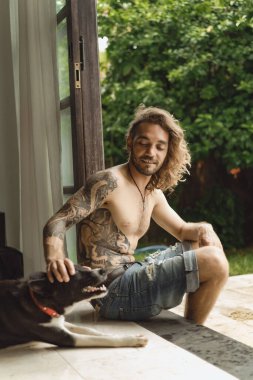 Uzun kıvırcık saçlı ve çıplak gövdeli genç bir adam. Bahçedeki sade bir ortamda bir adam bir köpekle oynuyor..