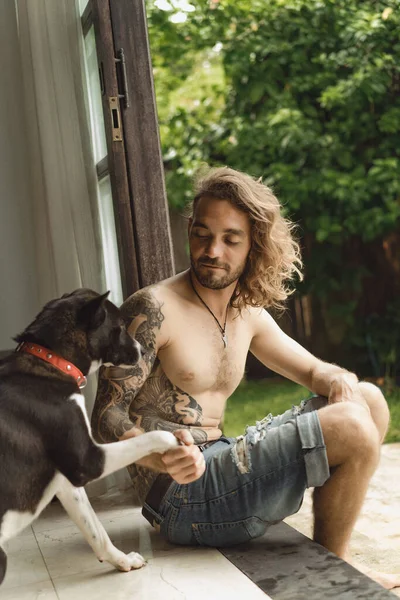 長い巻き毛と自宅で裸の胴を持つ若い男 庭の家庭的な雰囲気の中で犬と遊ぶ男 — ストック写真