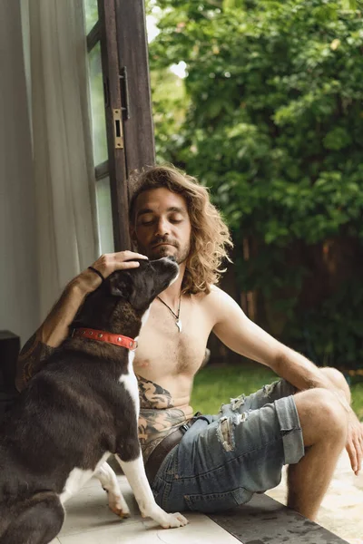 長い巻き毛と自宅で裸の胴を持つ若い男 庭の家庭的な雰囲気の中で犬と遊ぶ男 — ストック写真