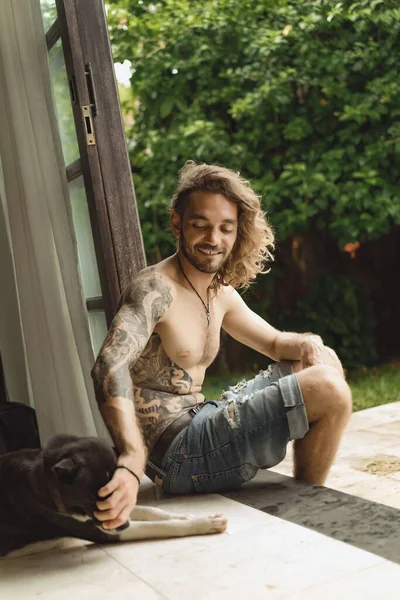 一个留着长长的卷发 赤身裸体的年轻人在家里 一个人在院子里和一只狗在舒适的气氛中玩耍 — 图库照片