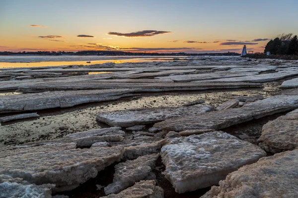 Frozen sea with floating ice blocks sheets against a distant red and white lighthouse during sunset in Charlottetown Prince Edward Island, Canada