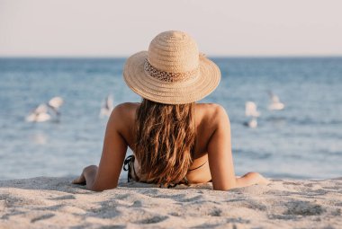 Young girl in a bathing suit and hat sitting on the sand on the beach, looking to the seagulls in the sea. Beautiful lady is facing the ocean with her back to the lens clipart