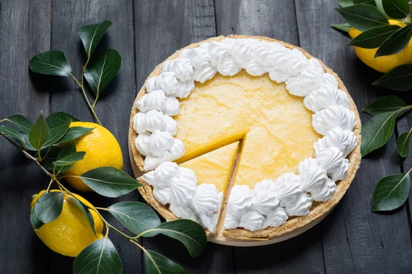 Homemade lemon pie with meringue, cutted piece, with fresh lemons on black wooden background, selective focus, horizontal orientation.