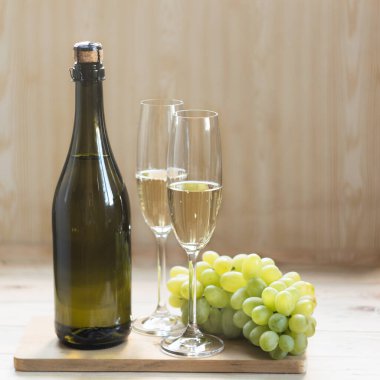 Bir şişe köpüklü şarap, iki kadeh köpüklü şarap ve hafif ahşap arka planda üzümler. Seçici odak.