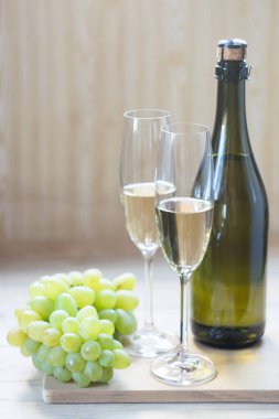 Bir şişe köpüklü şarap, iki kadeh köpüklü şarap ve hafif ahşap arka planda üzümler. Seçici odak.