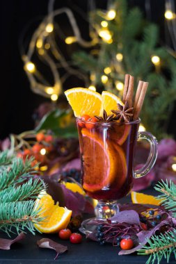 Portakal dilimli sıcak şarap ve baharatlar siyah ahşap masada Noel ışıkları ve arka planda ladin dalları. Seçici odak, yakın plan