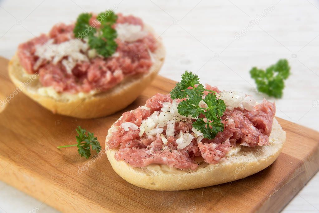 minced pork sausage, typical german mettwurst with onions and parsley garnish on a bun, kitchen board  on a white wooden table, close up with selected focus and narrow depth of field