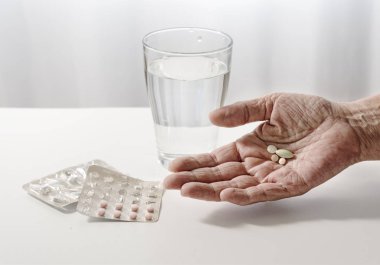elderly hand of a senior is holding pills, water glass and blister pack on the white table, light background with copy space, selected focus, narrow depth of field clipart