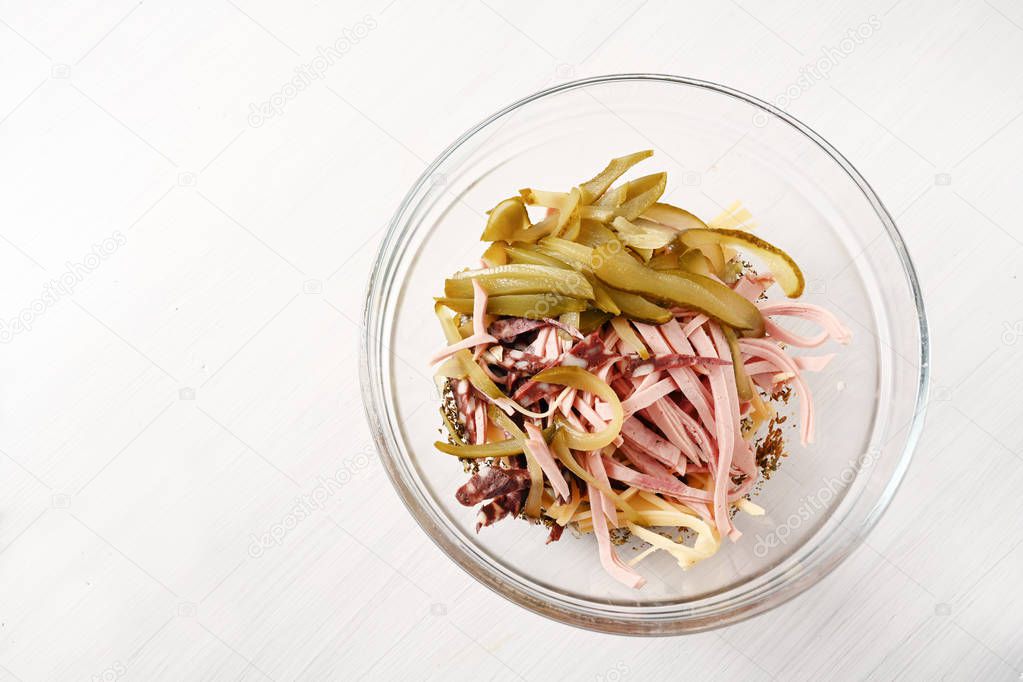 preparing sausage salad (german wurstsalat) in a glass bowl with onion, cheese and pickled gherkins, light background with copy space, high angle view from above