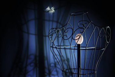 egg in a metal tailor doll like in a bird cage and a flying feather, surreal art concept for longing and loneliness, dark blue background with copy space clipart
