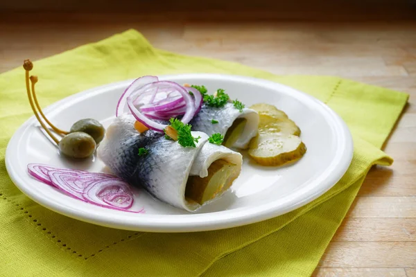 Rolled pickled herring, also called rollmops with red onions, gherkins and capers as a traditional hangover breakfast on a white plate, green napkin and a wooden table, copy space — Stock Photo, Image