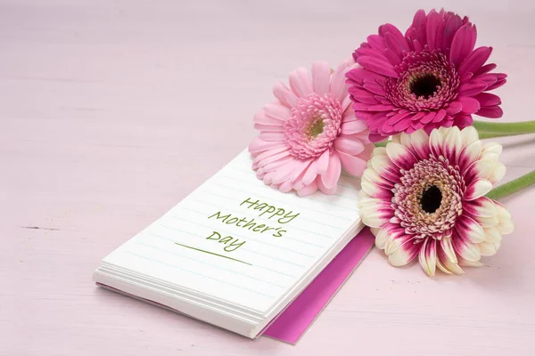 Three gerbera flowers lying on a writing pad, pastel pink colored background with copy space, text Happy Mother's Day — Stock Photo, Image