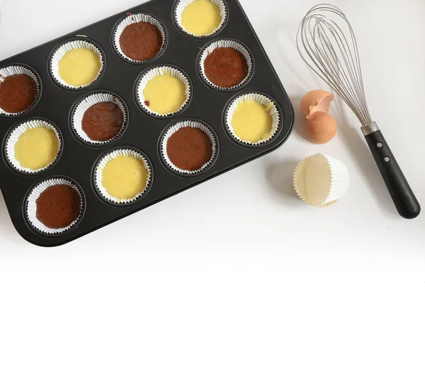 Muffin bakplaat gevuld met licht en donker taartdeeg voor cupcakes, whisk, eierschaal en papieren bekers, achtergrond vervaagt naar wit, grote Kopieer ruimte, groothoek aanzicht van bovenaf — Stockfoto