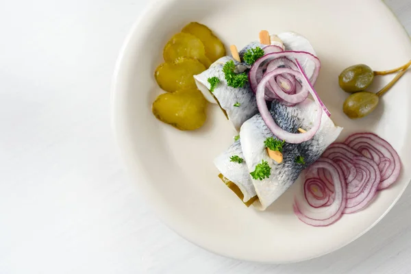 Rolou o arenque conservado em escabeche, também chamado rollmops com cebolas vermelhas, pepinos e alcaparras, um café da manhã de ressaca azedo em uma mesa branca, visão de alto ângulo de cima com espaço de cópia — Fotografia de Stock