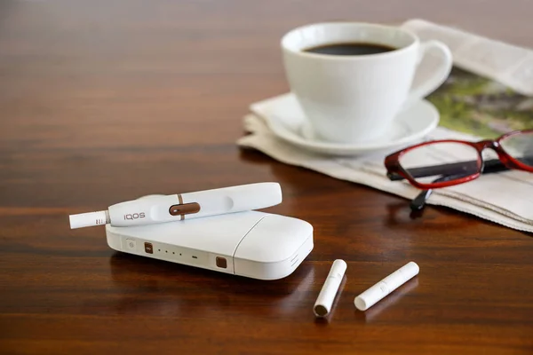 Rieps, Deutschland, 15. August 2019: iqos Heiztabakanlage, Kaffeetasse und Zeitung auf braunem Tisch, neue E-Zigarette mit Tabakstäbchen, die angeblich Schadensminderung bei Rauchern bewirkt, Kopierraum — Stockfoto