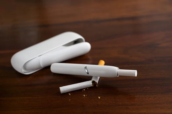 Biały system ogrzewania tytoniu łamie normalny papieros na brązowym stole, nowy rodzaj elektronicznego papierosa z pałkami tytoniu, który wytwarza aerozol nikotyny z mniejszą ilością środków toksycznych niż dym spalania i rzekomo powoduje redukcję szkód — Zdjęcie stockowe