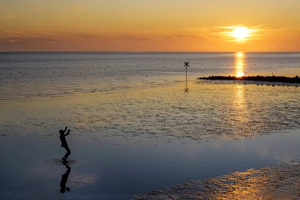 Niño pequeño como silueta está mirando al sol poniente en la playa del mar del norte en la marea baja, reflejos y gran espacio de copia —  Fotos de Stock
