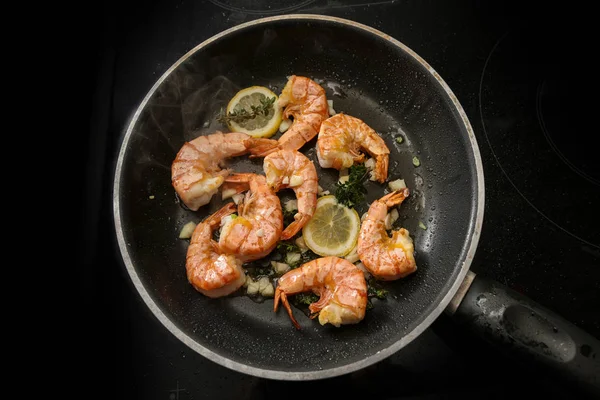 Tiger prawn shrimps are roasted in a black pan with garlic, lemon and herbs on a black stove, high angle view from above — Stock Photo, Image