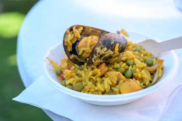 Spanyol utcai élelmiszer-, paella rizs tenger gyümölcsei és a kagyló egy fenntartható karton tálba és egy fából készült Villa egy fehér asztal, közelkép lövés — Stock Fotó