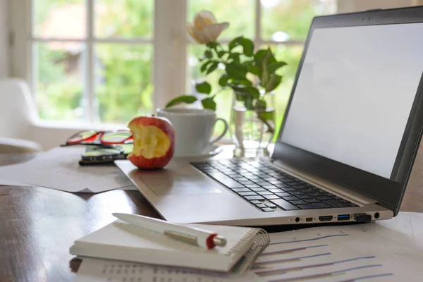 Home office espaço de trabalho para uma pequena empresa, na mesa um laptop com tela vazia, papéis, xícara de café, óculos e uma maçã mordida — Fotografia de Stock