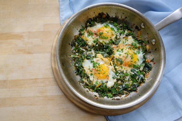 Pan with spinach and fried eggs, spices and herb garnish, protein-rich dish for low carb diet on a wooden table, copy space, high angle top view from above — Stock Photo, Image
