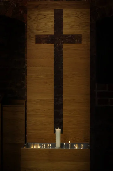 Velas grandes y pequeñas encendidas frente a una cruz simple moderna cortada de un plato de madera en la histórica iglesia de St. Georgen en Wismar, símbolo cristiano religioso sobre un fondo oscuro — Foto de Stock