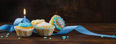 Koyu kahverengi bir zemin üzerinde mavi kurdeleli kekler ve yanan bir mum ile koyu kahverengi bir zemin üzerinde panoramik formatta fotokopi alanı olan kekler.