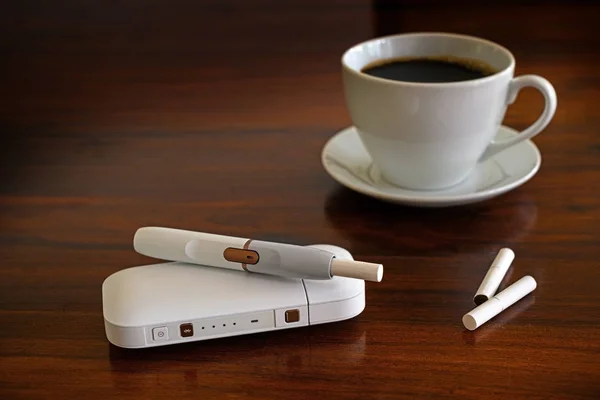 在棕色桌子上加热烟草系统和一杯咖啡，用烟草棒制成的电子香烟产生尼古丁气溶胶，据称比燃烧烟雾产生的有毒物质少，以减少吸烟者的伤害，复制空间 — 图库照片