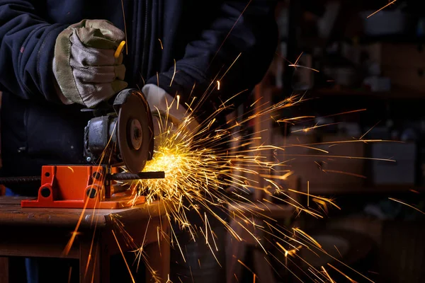 Manos de un artesano con guantes de trabajo cortando una barra de hierro con la amoladora angular eléctrica que rocía muchas chispas calientes en el taller oscuro, trabajo peligroso, espacio de copia —  Fotos de Stock