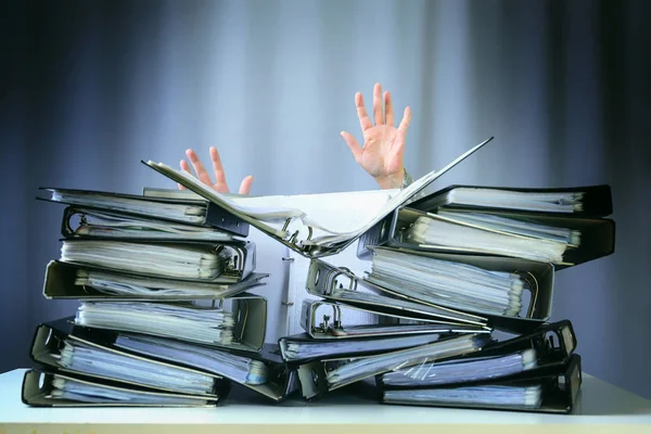 Upphöjda händer för en person som sjunker bakom staplar av ringpärmar på ett skrivbord, begreppet överdrivna krav och ökande arbete i näringslivet — Stockfoto