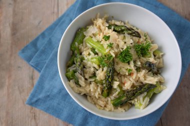 Beyaz bir kasede yeşil kuşkonmaz ve maydanozlu pilav, kopyalanmış ahşap üzerinde mavi peçete, yukarıdan yüksek açılı görüş, seçilmiş odak, dar alan derinliği.