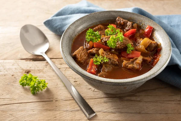 Fresh cooked goulash or stew from beef, onions and red bell pepper with parsley garnish in a bowl with spoon and a blue napkin on a rustic wooden table, selected focus, narrow depth of field