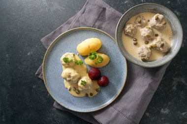 Koenigsberger Klopse or meatballs in white bechamel sauce with capers, potatoes and beetroot served in a bowl and on a gray blue plate, dark rustic background with copy space, high angle view from above clipart