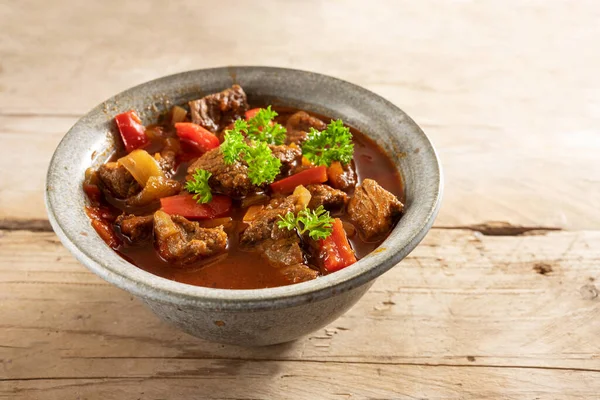 Fresh cooked goulash or stew from beef, onions and red bell pepper with parsley garnish in a bowl on a rustic wooden table, copy space, selected focus, narrow depth of field