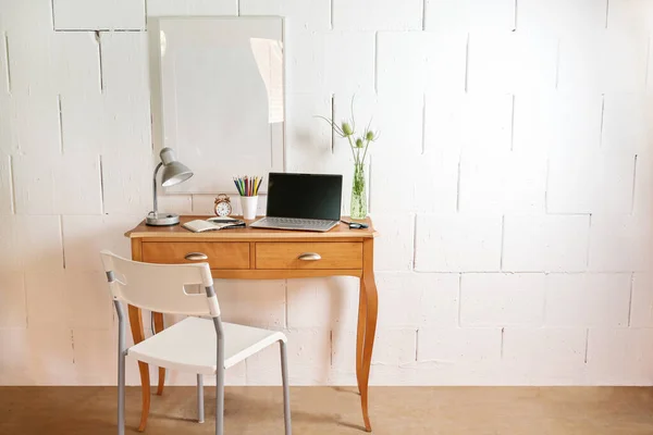 Small wooden table and chair as a tiny place for a home office with laptop, desk lamp and tools against a rough white wall, copy space, selected focus